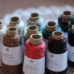 Peut inclure: Une collection de dix bobines de fil Creative Make It Paillette dans diverses couleurs, dont le rouge, le marron, le noir, le vert et la crème. Chaque bobine porte une étiquette en papier blanc avec du texte noir et des paillettes.