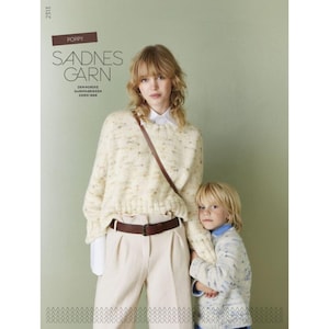 Op de afbeelding: Een vrouw en een kind dragen witte truien met een gespikkeld patroon. De vrouw draagt een bruine riem en een witte broek. De tekst "Sandnes Garn" is zichtbaar in de afbeelding.