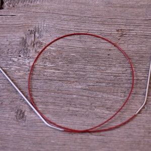 May include: A red circular knitting needle with two silver tips. The needle is lying on a wooden surface.