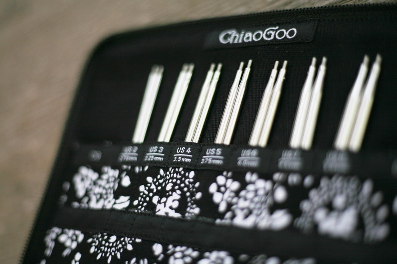 May include: A black zippered case with the text "ChiaoGoo" on the front. The case contains a set of silver knitting needles labeled with US sizes 2, 3, 4, 5, 6, 7, 8, 9, 10, and 11.