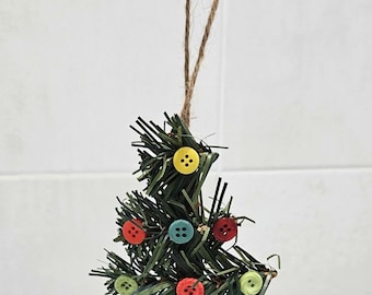 Può includere: Un ornamento per albero di Natale verde fatto di aghi di pino con bottoni colorati per decorazioni. L'ornamento è appeso a uno spago di iuta marrone.