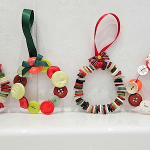 Puede incluir: Cuatro adornos navideños hechos a mano con botones coloridos, cada uno con un lazo de cinta para colgar. Los adornos tienen forma de coronas y presentan botones rojos, verdes, blancos y amarillos.