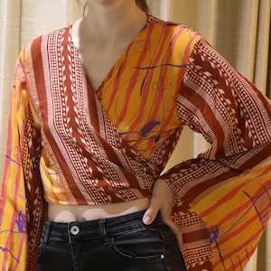 Pode incluir: Uma blusa transpassada de manga comprida com um padrão vibrante de riscas amarelas, cor de laranja e castanho escuro com motivos geométricos e florais. A peça tem um decote em V, bainha curta e mangas largas, combinada com calças de ganga pretas.