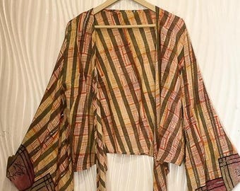 May include: A patterned kimono-style top with wide sleeves and a tie closure. The fabric features a striped design in shades of brown, orange, and yellow, with decorative panels at the sleeve ends. The top is hanging on a wooden hanger.