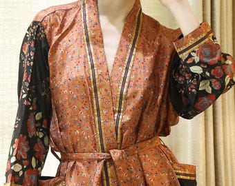 May include: A long-sleeved robe featuring a rust-orange base with a delicate floral pattern. The garment includes contrasting black sleeves and pockets adorned with a vibrant red and green floral print, finished with a matching tie belt at the waist.