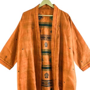 Può includere: Una giacca arancione in stile kimono con maniche a tre quarti è appesa a un'appendiabiti di legno. La giacca ha un davanti ampio e aperto, che rivela una fodera fantasia con accenti verdi e blu. Il tessuto esterno presenta piccoli dettagli ricamati scuri.