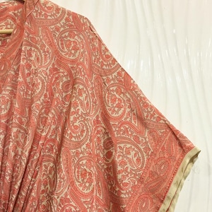 May include: A coral and cream kaftan featuring an intricate paisley pattern throughout. The garment has a V-neckline, wide kimono-style sleeves, and a gathered waist detail. The fabric displays a repeating floral and teardrop motif in a soft color palette.