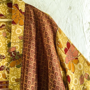 Könnte beinhalten: Nahaufnahme eines gelben Blumenkimonos, der an einem Holzbügel hängt. Der Kimono hat ein sich wiederholendes Muster aus großen orangefarbenen und rosa Blumen mit einem braunen geometrischen Muster auf den Ärmeln und am Kragen. Der Hintergrund ist eine weiße Wand.