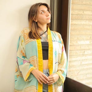 May include: A woman wearing a light green and yellow patterned kimono with a black crop top and denim shorts. The kimono has a yellow trim and a geometric design.