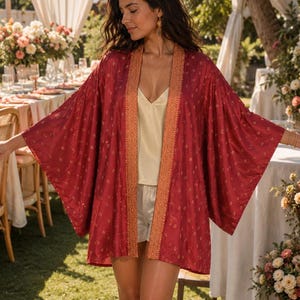 May include: A deep red kimono-style jacket featuring a repeating gold floral pattern and a wide, textured orange-gold border along the front opening. The garment has loose, wide sleeves and a straight hemline, worn over a cream-colored camisole.