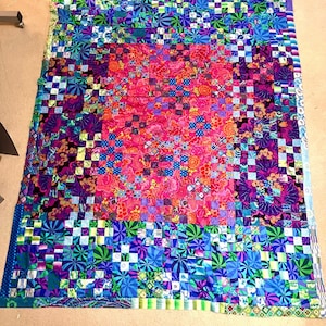 May include: A rectangular quilt featuring a central block of vibrant pink and orange floral fabric surrounded by a complex patchwork border. The border consists of small squares in shades of blue, purple, green, and teal with various geometric patterns.