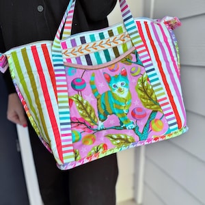 May include: A vibrant tote bag with vertical stripes in red, pink, green, and white. The central panel showcases a whimsical cat illustration on a pink background. The bag features a zip closure and matching striped handles.