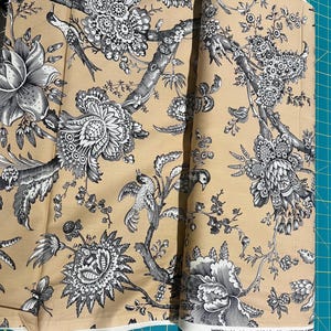 May include: Tan fabric with a black and white floral and bird pattern. The design features detailed illustrations of flowers, birds, and branches. The fabric is rolled up, showing the pattern's repeat.