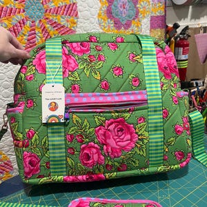 May include: A quilted green bag featuring a vibrant pink rose floral pattern, light blue polka dot trim, and green and yellow striped straps. A white tag attached to the front displays the text The LUCKY LUX with a circular graphic illustration.
