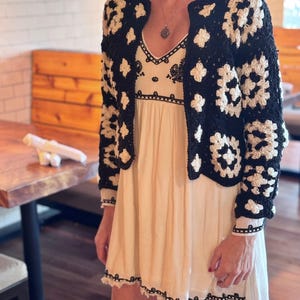 May include: A black and white crochet cardigan with a white crochet trim. The cardigan is worn over a white dress with black embroidery.