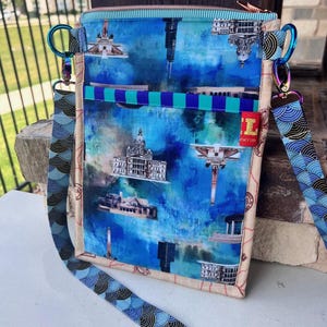 May include: A blue crossbody bag with a cityscape print, featuring buildings and a watercolor effect. The bag has a patterned strap and a zipper closure. The bag is approximately 8 inches wide and 10 inches tall.