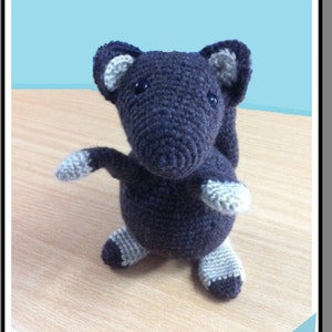 May include: A crocheted grey squirrel toy with white accents. The toy is standing on a wooden surface. The text "Silence the Squirrel" is displayed above the toy in a red banner.