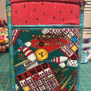 Puede incluir: Una bolsa rectangular de tela con una parte superior roja con estampado de sandía y un bolsillo frontal verde con temática de bolos. El bolsillo presenta bolos, una bola de bolos y las palabras "Welcome to Fabulous Bowling" y "Bingo".
