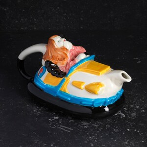 May include: A ceramic teapot shaped like a bumper car with a woman in a pink dress and brown hair driving. The teapot is painted in yellow, blue, white, and black.