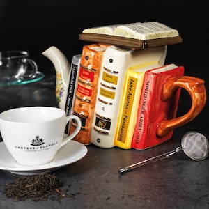 Può includere: Una teiera in ceramica a forma di pila di libri, con titoli come "Jane Austen - Persuasione" e "Northanger Abbey - 1818". La teiera presenta un design floreale e un manico marrone. In primo piano ci sono una tazza da tè e un piattino bianchi, insieme a un filtro per il tè.