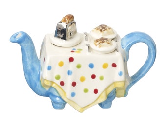 May include: A blue teapot shaped like a table with a white tablecloth with colorful polka dots. The teapot has a toaster and two pancakes on top.