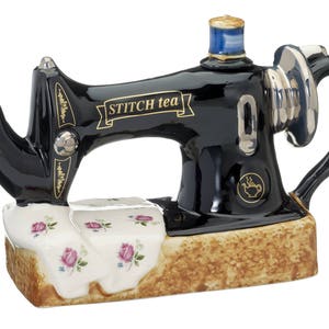 May include: Black ceramic teapot shaped like a vintage sewing machine with gold trim and the words "Stitch Tea" on the side. The teapot is sitting on a white and pink floral base.