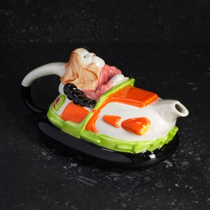 May include: A white ceramic teapot shaped like a hovercraft with a woman in a pink dress and brown hair driving it. The hovercraft is decorated with orange, green, and black details.