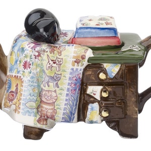 Könnte beinhalten: Eine Teekanne aus Keramik in Form einer Kommode mit einer Oberseite im Quiltmuster, einer schlafenden schwarzen Katze auf der Decke und einem Stapel Bücher auf der Kommode. Die Teekanne ist braun mit goldenen Akzenten und hat einen braunen Griff.
