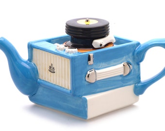 Puede incluir: Una tetera de cerámica azul y blanca con forma de tocadiscos vintage. La tetera tiene un disco negro en la parte superior y un asa plateada.