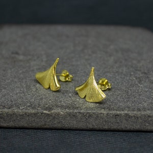 May include: A pair of gold-toned earrings shaped like ginkgo leaves. The earrings are on a gray stone surface.