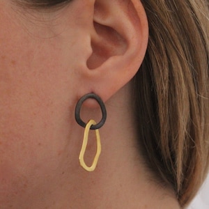 以下が含まれることがあります： ダークグレーの楕円形と、金色の不規則な涙の形が組み合わされたモダンなイヤリング。 イヤリングは耳に装着され、デザインと色のコントラストが際立っています。