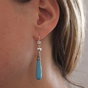 May include: Silver dangle earrings with a teardrop-shaped turquoise blue stone. The earrings have a floral design at the top and a detailed silver cone shape above the stone. The earrings are approximately 2.5 inches long.