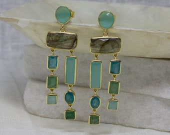 Puede incluir: Un par de pendientes colgantes de oro con un diseño en capas que presenta varios tonos de piedras preciosas verdes y azules. La parte superior de cada pendiente presenta una piedra preciosa redonda de color azul claro, seguida de una piedra preciosa rectangular de color gris, y luego una serie de piedras preciosas más pequeñas, rectangulares y ovaladas, en tonos de verde y azul.