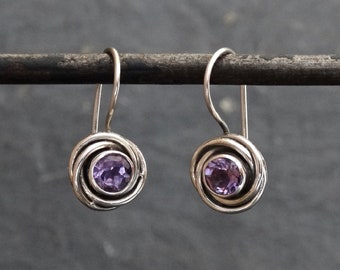 May include: A pair of silver earrings with a purple gemstone in the centre of each earring. The earrings are in the shape of a rose.