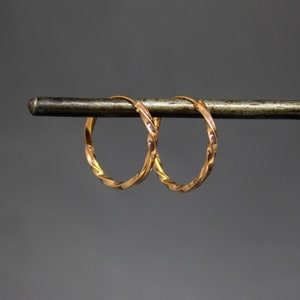 Op de afbeelding: Een paar goudkleurige oorringen met een gedraaid touwontwerp. De oorbellen worden getoond op een donkere metalen staaf tegen een grijze achtergrond. De hoepels zijn cirkelvormig en hebben een glanzend, reflecterend oppervlak.