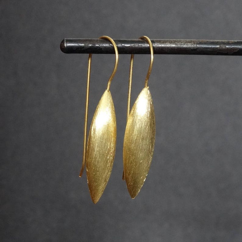 Puede incluir: Un par de pendientes de color dorado con acabado cepillado. Los pendientes tienen forma de hoja larga y estrecha y cuelgan de un gancho simple.