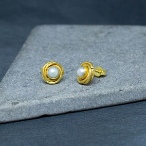May include: A pair of gold earrings with a white pearl in the center of each earring. The earrings are on a gray surface.