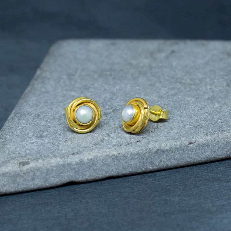 May include: A pair of gold earrings with a white pearl in the center of each earring. The earrings are on a gray surface.