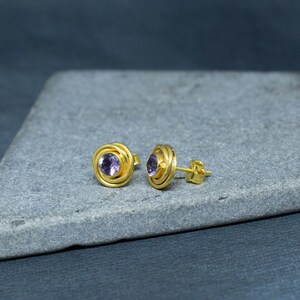 K&ouml;nnte beinhalten: Ein Paar goldene Ohrstecker mit einem violetten Amethyst-Edelstein in der Mitte jedes Ohrsteckers. Die Ohrstecker haben ein spiralf&ouml;rmiges Design um den Edelstein herum.
