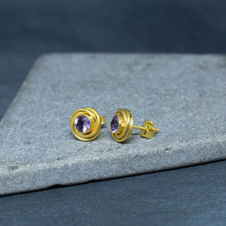 K&ouml;nnte beinhalten: Ein Paar goldene Ohrstecker mit einem violetten Amethyst-Edelstein in der Mitte jedes Ohrsteckers. Die Ohrstecker haben ein spiralf&ouml;rmiges Design um den Edelstein herum.