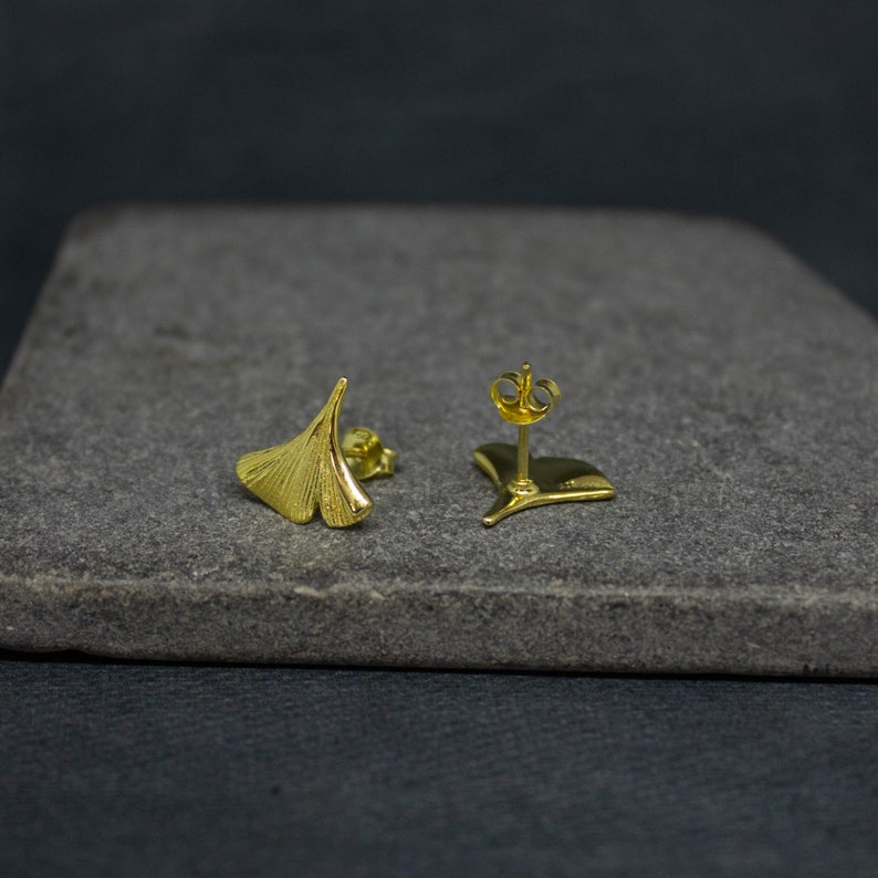 May include: A pair of gold-toned ginkgo leaf earrings. The earrings are small and delicate, and they have a simple, elegant design.