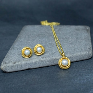 May include: A gold necklace and earrings set with a pearl in each piece. The necklace has a delicate chain and a pendant with a pearl in the center. The earrings are stud earrings with a pearl in the center.