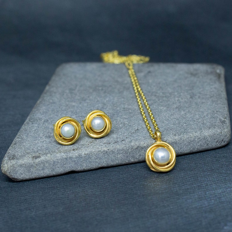 May include: A gold necklace and earrings set with a pearl in each piece. The necklace has a delicate chain and a pendant with a pearl in the center. The earrings are stud earrings with a pearl in the center.