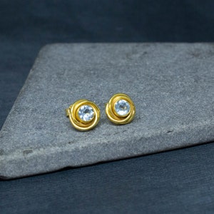 May include: A pair of gold earrings with a clear round gemstone in the center of each earring.