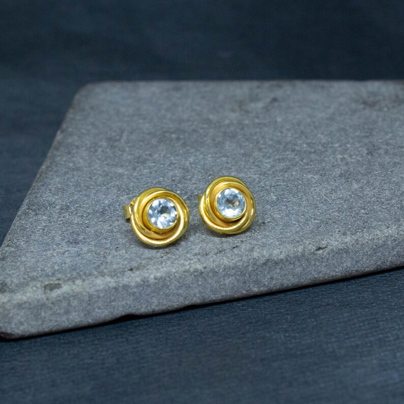 May include: A pair of gold earrings with a clear round gemstone in the center of each earring.