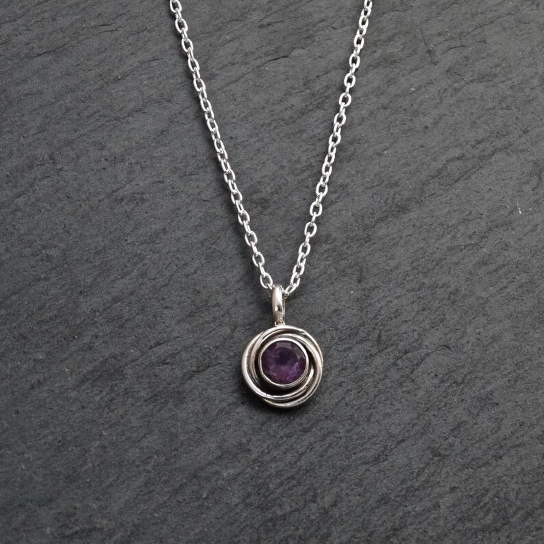 K&ouml;nnte beinhalten: Eine silberne Kettenkette mit einem Anh&auml;nger, der einen violetten Amethyst-Edelstein in einem silbernen Wirbeldesign zeigt.