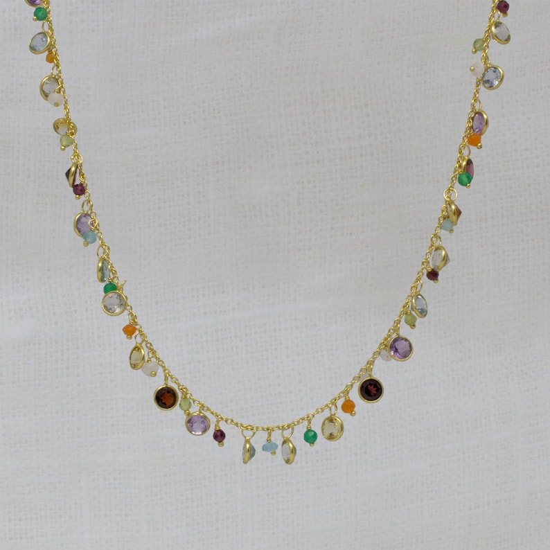 May include: A gold chain necklace with a variety of colorful gemstones hanging from it. The gemstones are in different shapes and sizes, and they include amethyst, aquamarine, citrine, garnet, peridot, and rose quartz.