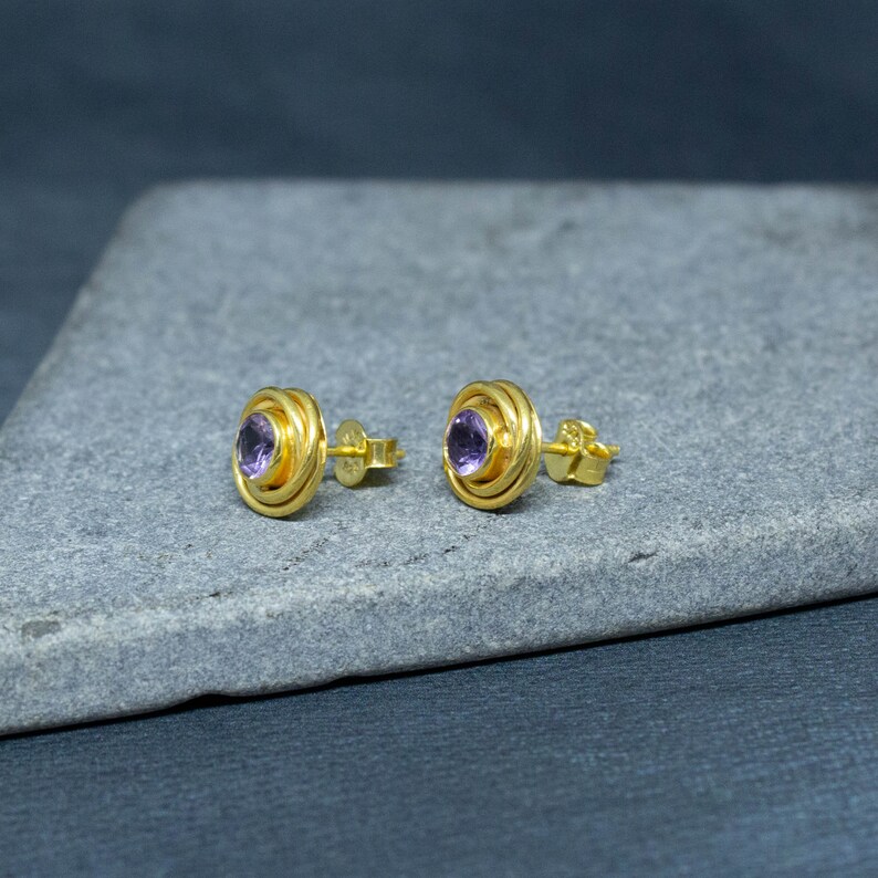 K&ouml;nnte beinhalten: Ein Paar goldene Ohrringe mit einem violetten Edelstein in der Mitte. Die Ohrringe haben ein kreisf&ouml;rmiges Design mit mehreren Ringen um den Edelstein.