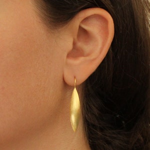 Puede incluir: Pendientes de hoja de oro con forma ovalada larga y estrecha. Los pendientes cuelgan de la oreja de una persona.
