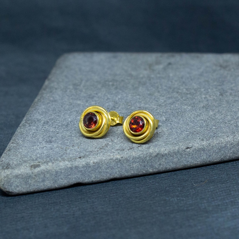 Op de afbeelding: Een paar gouden oorbellen met rode edelsteen accenten. De oorbellen hebben een cirkelvormig ontwerp met meerdere gouden ringen.